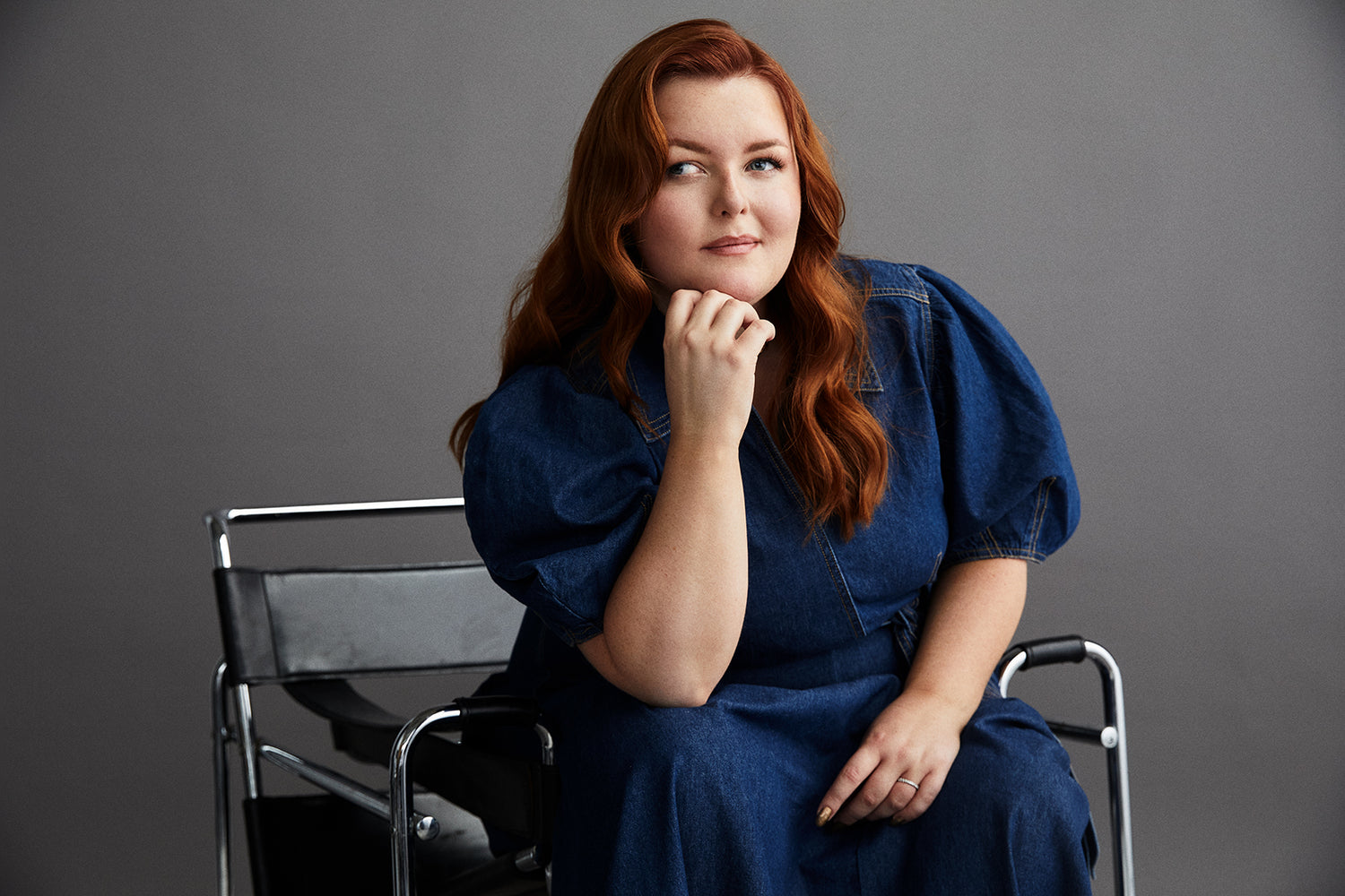 Ginger haired Lucy Edwards wears a denim dress and sits on a chair thinking against a grey background